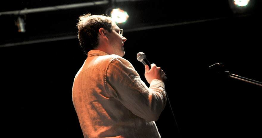 Lutz Schmitt on a stage with a microphone. Background dark, white spotlights on him.